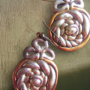 May include: A pair of handcrafted silver-toned earrings. Each earring features a bow-shaped top and a circular, woven design with a copper-colored border. The earrings are displayed against a textured, olive-green background.