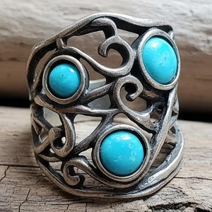 May include: A silver-toned ring with an openwork design featuring three round, bright turquoise stones. The ring has a detailed, swirling pattern and a vintage aesthetic, suitable for jewelry collections.
