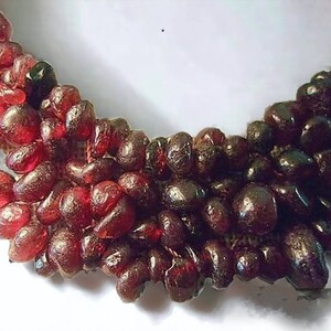 May include: A close-up of a necklace made of dark red, irregularly shaped beads. The beads have a glossy, translucent appearance, with variations in color from lighter red to deep burgundy. The necklace is arranged in multiple strands.
