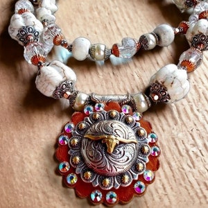May include: A handcrafted necklace featuring white, marbled beads and clear crystal accents. The pendant is a silver-toned metal with a gold bull design, surrounded by a red leather backing and iridescent crystals. The necklace is designed with a rustic, bohemian style.