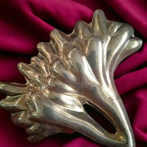 Puede incluir: Una escultura de metal plateado y abstracta con un diseño orgánico y fluido. La escultura tiene una superficie texturizada y está colocada sobre un fondo de tela rojo oscuro. La pieza tiene una forma artística única.