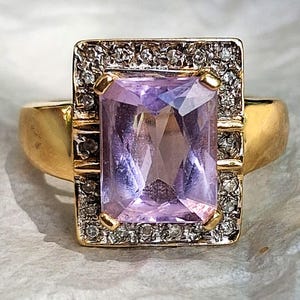 May include: A gold ring featuring a large, rectangular, light purple gemstone. The gemstone is surrounded by a square frame of smaller, clear stones. The ring's band is a simple, polished gold.