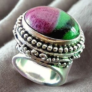 May include: A silver ring with an oval-shaped gemstone. The gemstone is divided into two colors: a deep magenta and a vibrant green. The ring has a detailed, beaded design around the stone and a smooth, polished band. The ring is set against a soft, gray background.