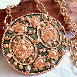 May include: A large circular gold-tone pendant necklace featuring a textured green enamel background. The pendant is decorated with multiple raised gold-tone circular medallions and small metallic spheres, suspended from a thick gold-tone chain.