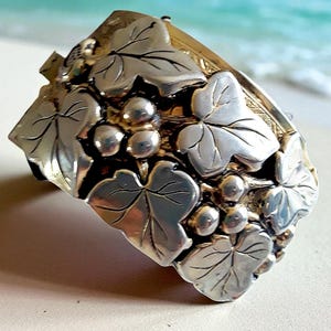 May include: Silver-toned cuff bracelet featuring a grape and leaf design. The bracelet has detailed leaf and grape cluster embellishments. The metal has a polished finish, reflecting light. The clasp is visible on the side. The bracelet is a piece of jewelry.
