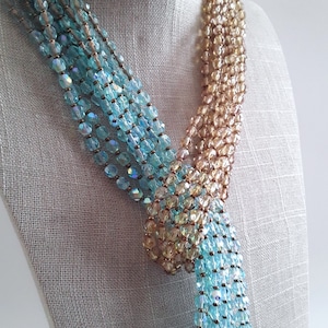 May include: A multi-strand necklace featuring faceted beads in shades of light blue and champagne gold. The strands are gathered and knotted together, creating a layered effect with iridescent finishes on the blue beads and a metallic sheen.