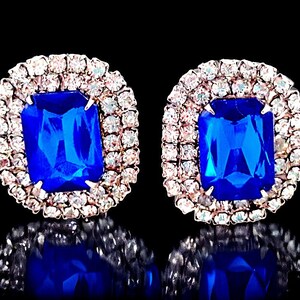 Puede incluir: Un par de pendientes de piedras azules con engaste plateado. Las piedras preciosas están rodeadas por un halo de piedras pequeñas transparentes.