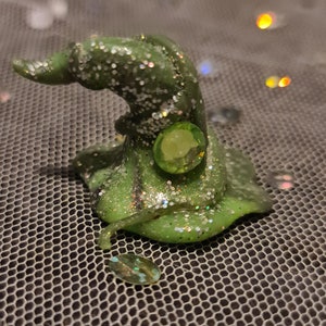 May include: A green glitter witch hat with a green gem on top. The hat is sitting on a mesh surface.