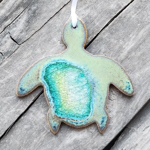 May include: A green ceramic turtle ornament with a blue and green glaze. The ornament is hanging from a white ribbon.
