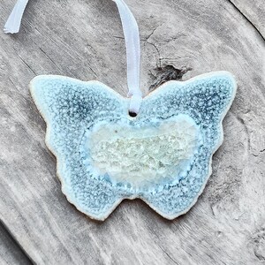 May include: A light blue ceramic butterfly ornament with a white and green glaze. The butterfly is hanging from a white ribbon.