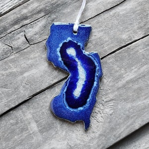 May include: Blue and white ceramic ornament shaped like the state of New Jersey. The ornament has a white ribbon attached for hanging.