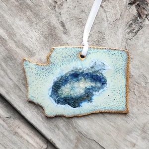 May include: A ceramic ornament shaped like the state of Washington with a blue and white agate design. The ornament is hanging from a white ribbon against a wooden background.