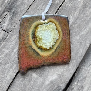 May include: A ceramic ornament shaped like the state of New Mexico with a heart-shaped design in the center. The ornament is brown, red, and yellow with a white heart.