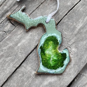 May include: A green and white ceramic ornament shaped like the state of Michigan. The ornament has a green glaze and a white outline. It is hanging from a white ribbon.