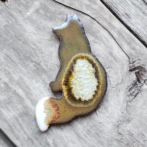 May include: A ceramic fox figurine with a brown and yellow glaze. The fox is sitting with its tail curled around its body. The fox's body has a white center with a textured design.