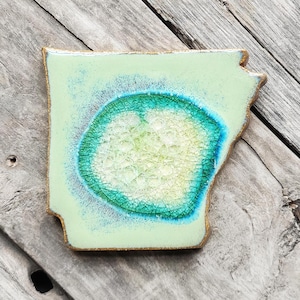 May include: A ceramic state-shaped coaster in the shape of Arkansas. The coaster is light green with a light blue and green circle in the center. The circle has a cracked glass-like texture.