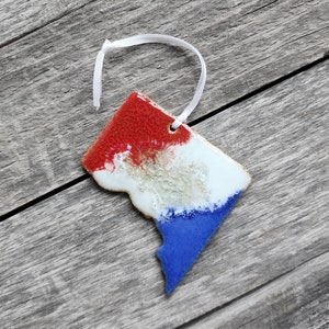 May include: A ceramic ornament shaped like the District of Columbia, painted in red, white, and blue with a white ribbon.