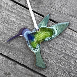 May include: A handcrafted hummingbird ornament with a white ribbon for hanging. The bird features a long, purple beak, blue and green body, and a white outline. The ornament is set against a weathered wooden background.