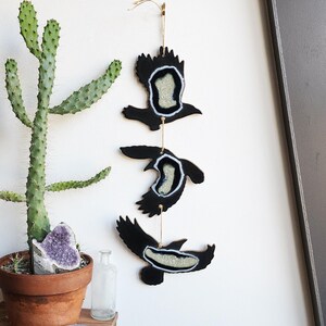 May include: A wall hanging featuring three black bird silhouettes with agate slices in the center. The birds are suspended from a gold string and are arranged vertically.