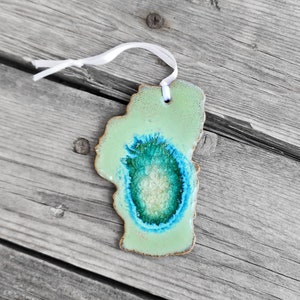 May include: A green ceramic ornament shaped like a lake with a blue and green swirl in the center. The ornament has a white string attached to it.