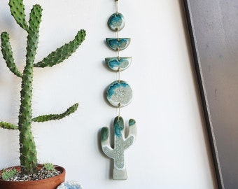 May include: A ceramic wall hanging with a cactus design and a series of blue and green circles and half-circles. The hanging is attached to a white wall with a string.