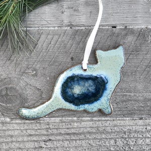 May include: A ceramic cat ornament with a blue and green glaze. The ornament is shaped like a cat sitting down and has a white ribbon attached to the top.