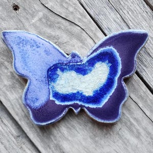May include: A ceramic butterfly wall decor with a blue and purple glaze. The butterfly has a white center with a crackled glaze.