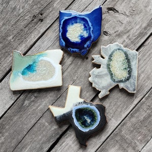 May include: Four ceramic state-shaped coasters with a geode design. The coasters are in the shapes of Oregon, Ohio, Texas, and Pennsylvania. The geode designs are in shades of blue, green, and white.