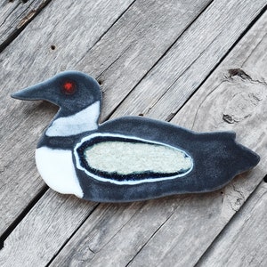 May include: A ceramic loon figurine with a black body, white accents, and a red eye. The bird-shaped ornament is set against a weathered wooden background. The loon has a light green oval shape on its body.