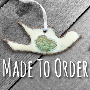 May include: White ceramic dove ornament with a green and brown glaze. The dove is hanging from a white ribbon and the text "MADE TO ORDER" is printed below the dove.