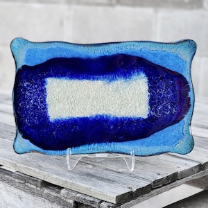 May include: A rectangular glass tray with wavy edges featuring a gradient of colors. The center is a textured white rectangle surrounded by a deep royal blue band, transitioning into a lighter sky blue border at the outer edges of the piece.