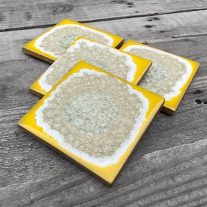 May include: Four square coasters with a yellow border and a white and beige center, resembling a geode. The coasters are arranged on a weathered wooden surface. The design is a unique and decorative addition to any home.