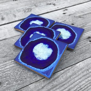 May include: Four blue and white ceramic coasters with a geode design. The coasters have a dark blue background with a white center that looks like a crystal.
