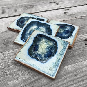 May include: Four square ceramic coasters with a blue and white agate design. The coasters have a gold rim and are stacked on top of each other.