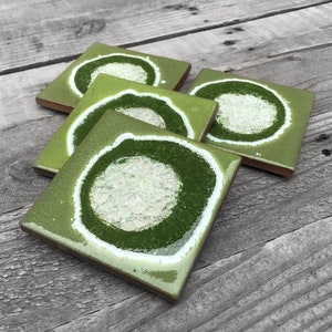 May include: Four green ceramic coasters with a white ring and a white center filled with small pieces of glass. The coasters are stacked on top of each other.