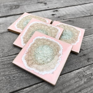 May include: Set of four square coasters with a pink glossy finish. Each coaster features a central design with a light brown, textured pattern, surrounded by a white border. The coasters are arranged on a weathered wooden surface.