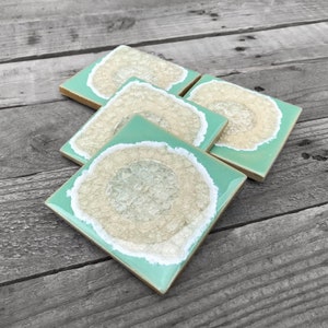 May include: Set of four square ceramic coasters with a light green background and a white outline. Each coaster features a beige agate-like design with a white center.