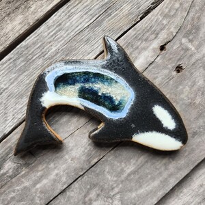 May include: A black and white ceramic orca whale wall hanging with a blue and green glaze. The orca has a white underbelly and a black back with a blue and green glaze inside the orca's body.