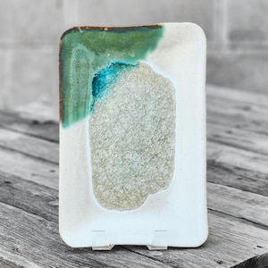 May include: A white ceramic rectangular dish with a green and blue glaze. The glaze has a textured, organic shape in the center of the dish.