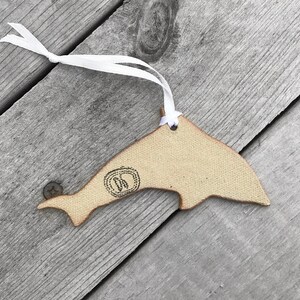 K&ouml;nnte beinhalten: Beige delfinf&ouml;rmiger Ornament mit wei&szlig;em Band zum Aufh&auml;ngen. Das Ornament hat ein gestempeltes Design in der N&auml;he des Schwanzes. Der Hintergrund besteht aus verwitterten, grauen Holzplanken.