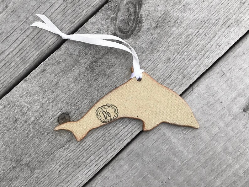 K&ouml;nnte beinhalten: Beige delfinf&ouml;rmiger Ornament mit wei&szlig;em Band zum Aufh&auml;ngen. Das Ornament hat ein gestempeltes Design in der N&auml;he des Schwanzes. Der Hintergrund besteht aus verwitterten, grauen Holzplanken.