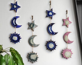 May include: Three ceramic wall hangings with a celestial theme. Each hanging features a star, moon, and sun with a blue, green, and pink glaze. The sun and moon have a white center with a blue, green, or pink outer ring.