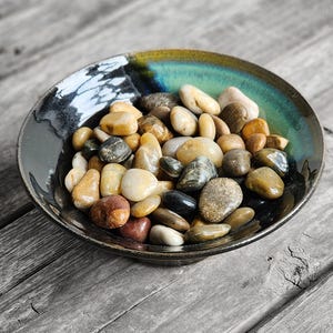May include: A ceramic bowl with a dark rim and a teal and gold interior holds a collection of smooth, colorful stones. The stones vary in size and color, including shades of brown, beige, gray, and black. The bowl sits on a weathered wooden surface.