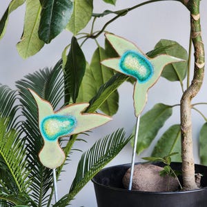 May include: Two green ceramic hummingbird garden stakes with blue and green glitter accents. The stakes are inserted into a potted plant.