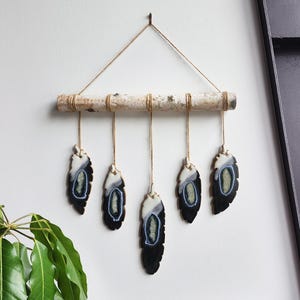 May include: A wall hanging made of a birch branch with five agate slices hanging from it. The agate slices are black and white with a blue and green center. The slices are suspended from the branch with twine.