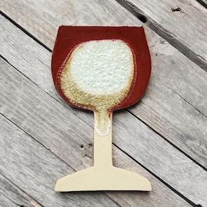 May include: A decorative wine glass-shaped object with a red rim and a white and gold textured center. The stem and base are a light beige color. The object is made of wood or ceramic.