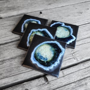 May include: Four black square coasters with a white and blue agate design. The coasters have a dark background with a white and blue agate design in the center.