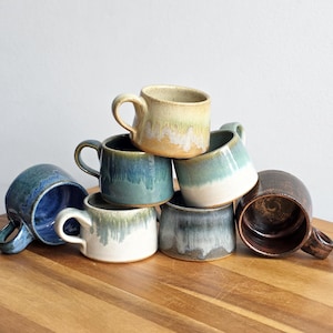 May include: A collection of seven ceramic mugs stacked in a pyramid formation. Each mug features a unique reactive glaze finish with varying color combinations including shades of blue, green, cream, white, and dark brown, all with a glossy texture.