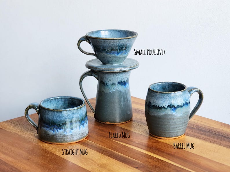 May include: A collection of handmade ceramic mugs and a small pour-over coffee maker. The mugs include a straight mug, flared mug, and barrel mug, all with a blue and gray glaze. The text on the image reads "Small Pour Over", "Flared Mug", "Straight Mug", and "Barrel Mug".