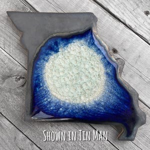 May include: A ceramic dish in the shape of the state of Missouri, with a dark gray border and a blue and white center. The text "SHOWN IN TIN MAN" is at the bottom. The dish is on a wooden surface.